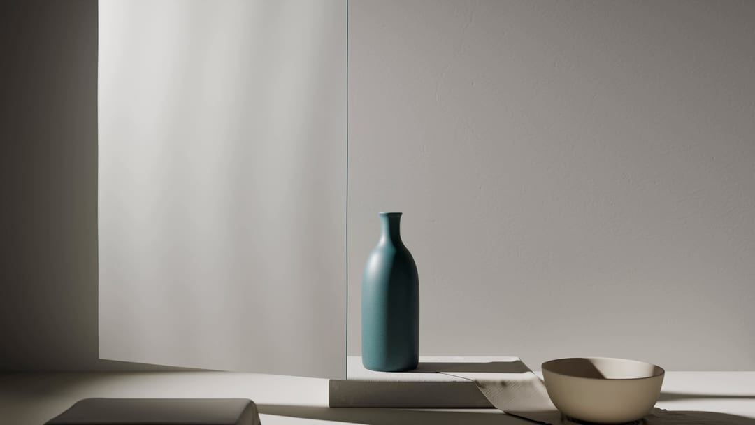 A matte, petrol-colored vessel stands on a low plinth in front of a pale wall, a pale bowl beside it. Light falls from the left onto a length of paper.